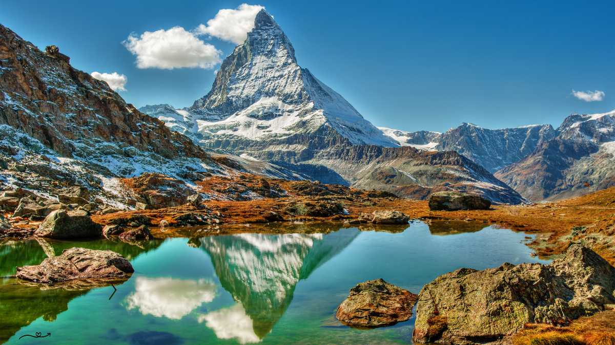 A majestic view of the Matterhorn under a cloudy sky, with swirling mist partially obscuring the sharp peak, surrounded by rocky ridges and snow, with a tranquil Alpine meadow below.